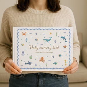 May include: A white baby memory book with a blue scalloped border and sea creature illustrations. The cover features a whale, dolphin, fish, and other marine life. The text "Baby memory book" and a name are printed on the cover.