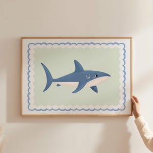 May include: A framed print featuring a cartoon shark in shades of blue and white. The shark is set against a light green background with a scalloped border. The frame is made of light brown wood.