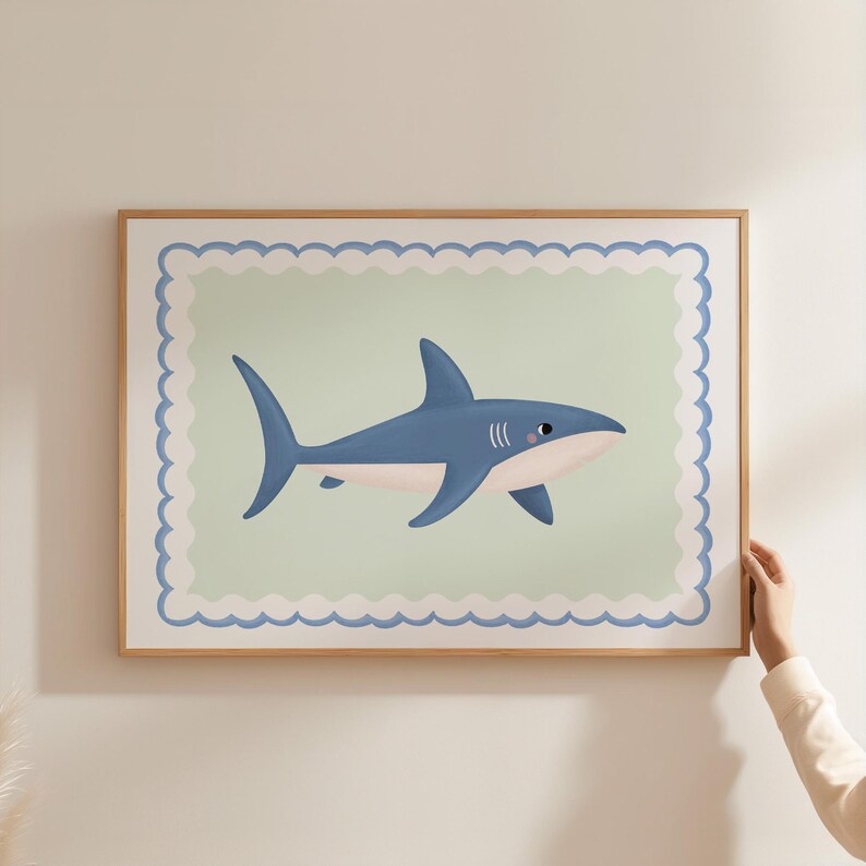 May include: A framed print featuring a cartoon shark in shades of blue and white. The shark is set against a light green background with a scalloped border. The frame is made of light brown wood.