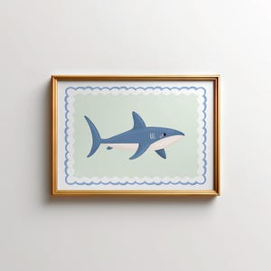 May include: A framed print of a cartoon shark. The shark is depicted in blue with white details and a pink mouth, set against a pale green backdrop with a scalloped edge. The frame is gold-coloured.
