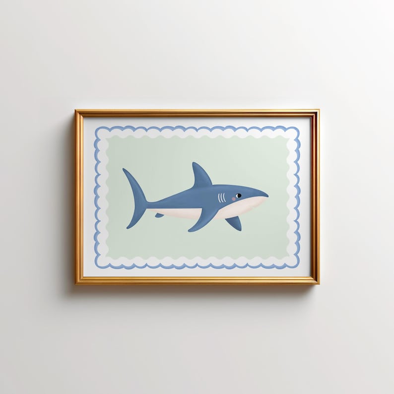May include: A framed print of a cartoon shark. The shark is depicted in blue with white details and a pink mouth, set against a pale green backdrop with a scalloped edge. The frame is gold-coloured.