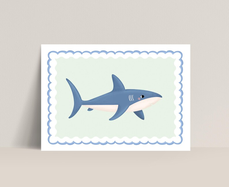 May include: A framed print featuring a cartoon shark. The shark is blue with a white belly and a small pink blush on its cheek. It is set against a light green background within a blue scalloped border.