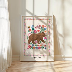 May include: A framed art print featuring a brown bear walking through a field of colorful flowers, butterflies, and a bee. The print has a decorative border and the words "little bear" at the bottom.