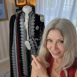 May include: A black coat with silver trim and a red interior, displayed on a mannequin. The coat features ornate detailing, including silver lace and a decorative emblem. The mannequin also has a white ruffled collar and cuffs. A framed vintage poster is visible in the background.