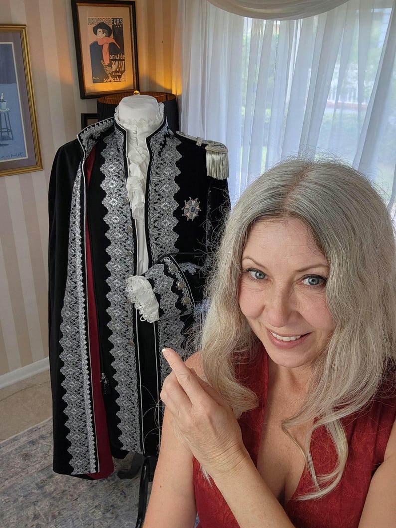 May include: A black coat with silver trim and a red interior, displayed on a mannequin. The coat features ornate detailing, including silver lace and a decorative emblem. The mannequin also has a white ruffled collar and cuffs. A framed vintage poster is visible in the background.