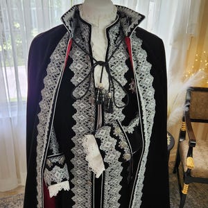 May include: A black velvet cape with white lace trim and a red lining. The cape is part of a costume, with a matching jacket and cuffs. The costume is displayed on a mannequin, with a white shirt and a high collar.