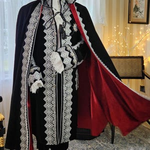 May include: A black velvet costume with white lace trim and a red satin lining. The costume includes a long cape, jacket, and ruffled sleeves. The outfit is displayed on a black mannequin stand.