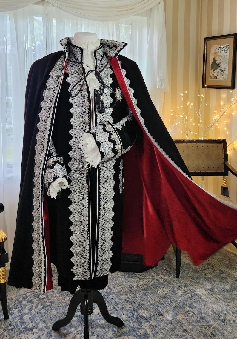 May include: A black velvet costume with white lace trim and a red satin lining. The costume includes a long cape, jacket, and ruffled sleeves. The outfit is displayed on a black mannequin stand.