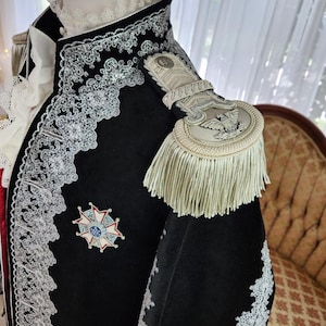 May include: A black velvet jacket with ornate silver lace trim and a decorative emblem. The jacket features a high collar, epaulettes with fringe, and a white ruffled undershirt. The jacket is part of a historical costume.