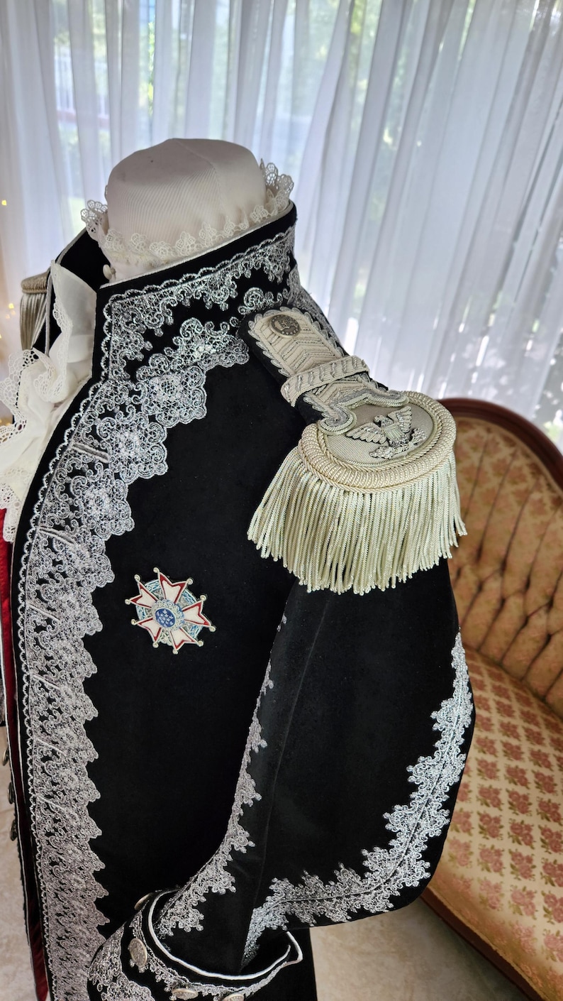 May include: A black velvet jacket with ornate silver lace trim and a decorative emblem. The jacket features a high collar, epaulettes with fringe, and a white ruffled undershirt. The jacket is part of a historical costume.