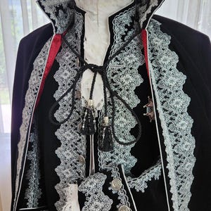 May include: A black velvet costume with white lace trim and red accents. The costume features a high collar, decorative buttons, and a cape. The intricate lace detailing and silver accents add to the formal appearance. The costume is displayed on a mannequin.