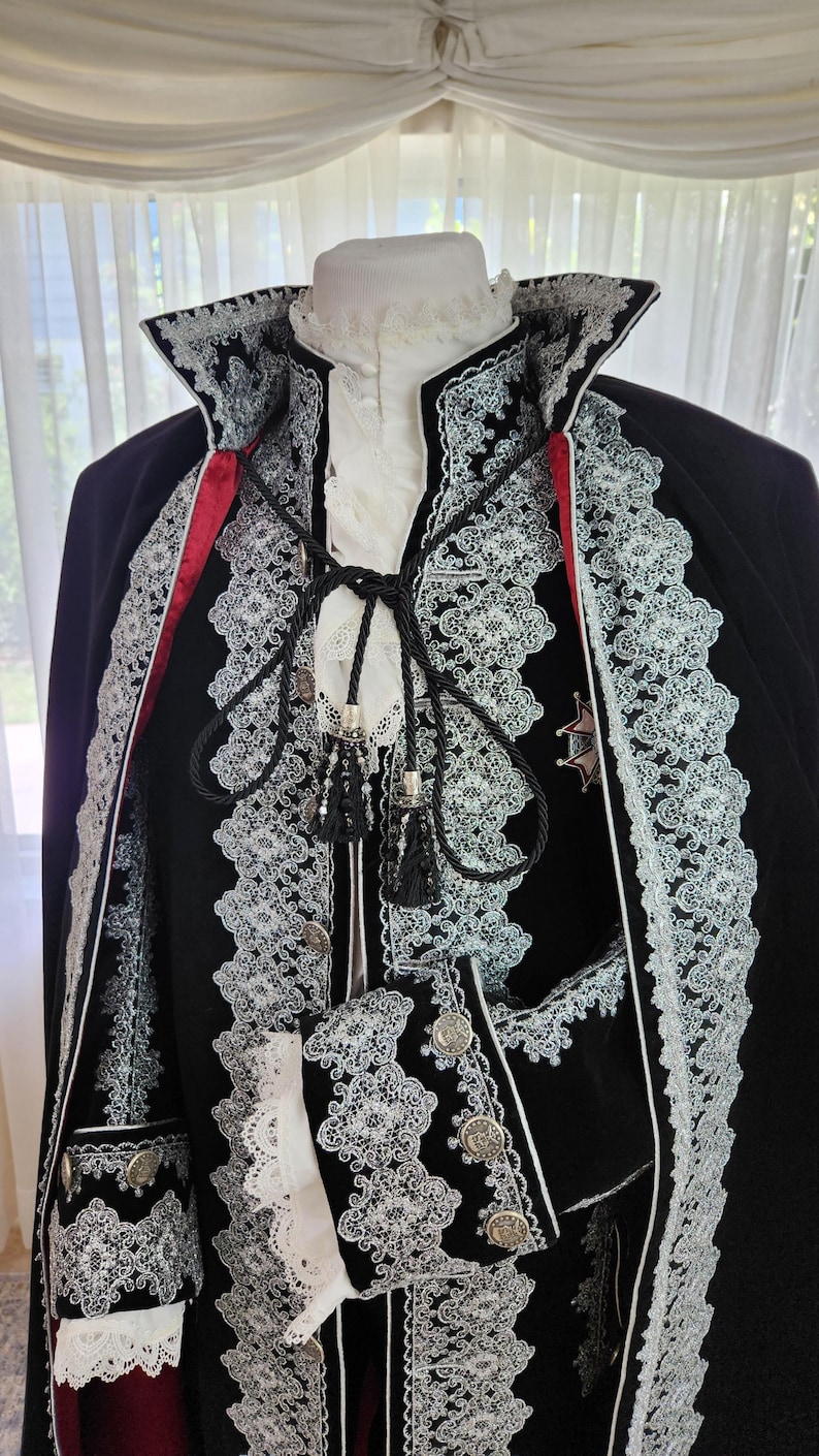 May include: A black velvet costume with white lace trim and red accents. The costume features a high collar, decorative buttons, and a cape. The intricate lace detailing and silver accents add to the formal appearance. The costume is displayed on a mannequin.