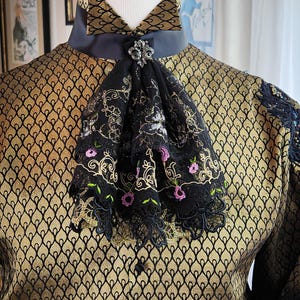 May include: A black and gold brocade shirt with a black lace trim and a black lace bow tie with gold embroidery and a small jeweled pin.