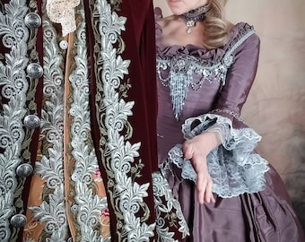 May include: A woman in a purple gown with a silver and white lace trim stands next to a man in a burgundy velvet jacket with silver embroidery. The woman is wearing a silver necklace and a brown feathered hairpiece.