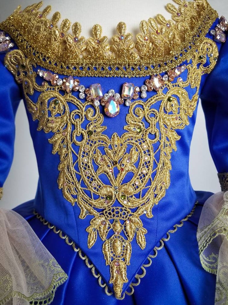 K&ouml;nnte beinhalten: Ein blaues Satin-Kleid mit goldener Spitze und Kristallverzierungen. Das Kleid hat einen herzf&ouml;rmigen Ausschnitt und ein tailliertes Oberteil. Der Rock ist voll und flie&szlig;end.