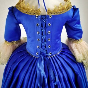 K&ouml;nnte beinhalten: Ein k&ouml;niglich blaues Satin-Kleid mit goldener Spitzenborte und einer blauen Schleife im R&uuml;cken. Das Kleid hat ein tailliertes Oberteil und einen vollen Rock.