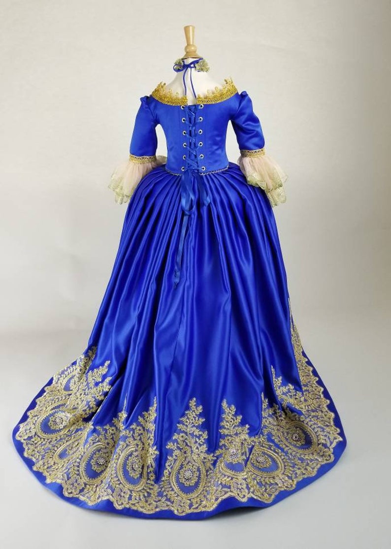 K&ouml;nnte beinhalten: Ein k&ouml;niglich blaues Satin-Ballkleid mit goldener Spitzenverzierung und einem Schn&uuml;rr&uuml;cken. Das Kleid hat einen vollen Rock und ein tailliertes Oberteil.