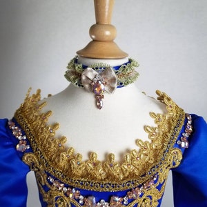 K&ouml;nnte beinhalten: Ein blaues Satin-Kleid mit goldener Spitzenborte und einem mit Juwelen besetzten Ausschnitt. Das Kleid hat eine passende blaue und gr&uuml;ne Schleifenkette mit einem mit Juwelen besetzten Bogen.