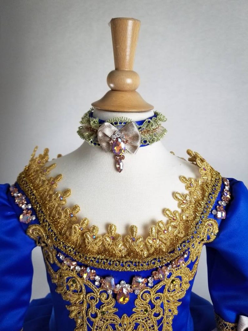 K&ouml;nnte beinhalten: Ein blaues Satin-Kleid mit goldener Spitzenborte und einem mit Juwelen besetzten Ausschnitt. Das Kleid hat eine passende blaue und gr&uuml;ne Schleifenkette mit einem mit Juwelen besetzten Bogen.