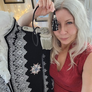 May include: A black military-style jacket with silver trim and a decorative emblem. The jacket features epaulettes with gold fringe and intricate silver lace detailing. A black tassel with beads is held up in front of the jacket.