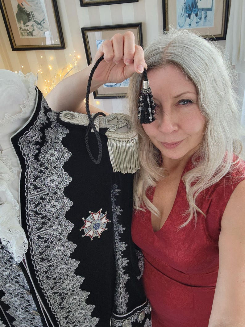 May include: A black military-style jacket with silver trim and a decorative emblem. The jacket features epaulettes with gold fringe and intricate silver lace detailing. A black tassel with beads is held up in front of the jacket.