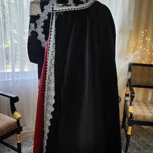 May include: A black cloak with a red lining and white lace trim, displayed on a stand. The garment features ornate embroidery and a white collar. A military-style jacket with gold epaulettes is visible.