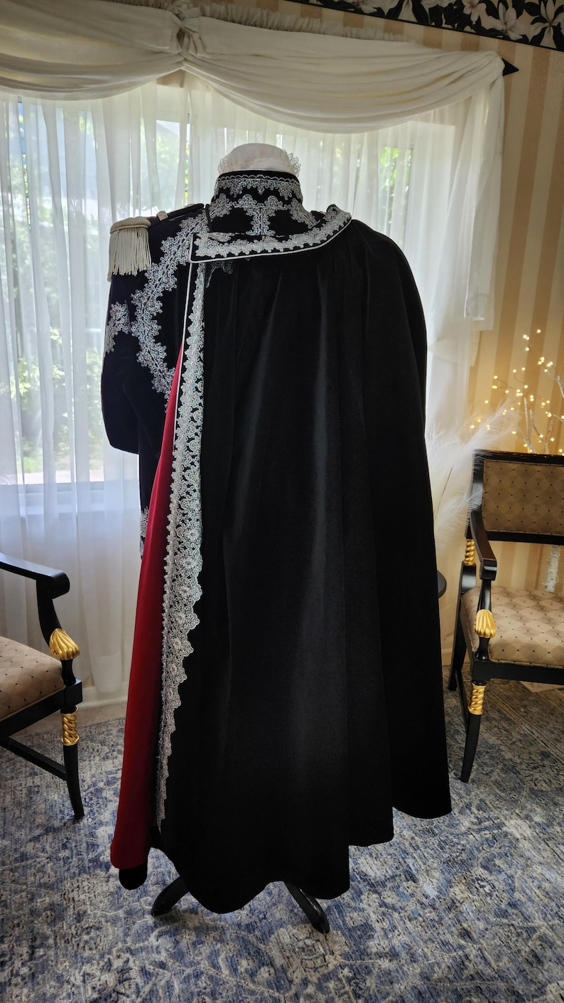 May include: A black cloak with a red lining and white lace trim, displayed on a stand. The garment features ornate embroidery and a white collar. A military-style jacket with gold epaulettes is visible.