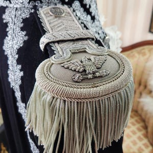 May include: Close-up of a black velvet garment with ornate silver embroidery and a detailed epaulette. The epaulette features an eagle emblem and silver fringe. The garment has a white ruffled collar.