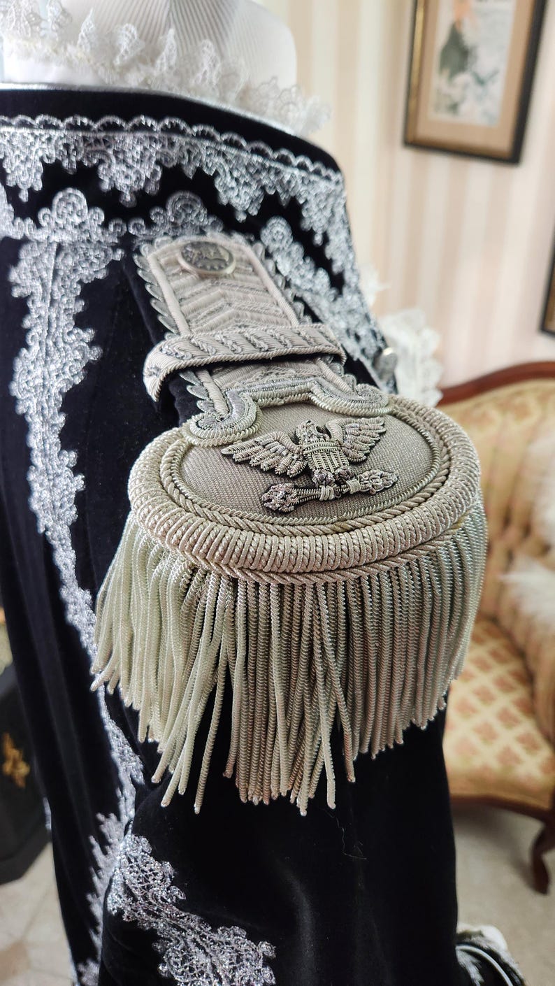 May include: Close-up of a black velvet garment with ornate silver embroidery and a detailed epaulette. The epaulette features an eagle emblem and silver fringe. The garment has a white ruffled collar.