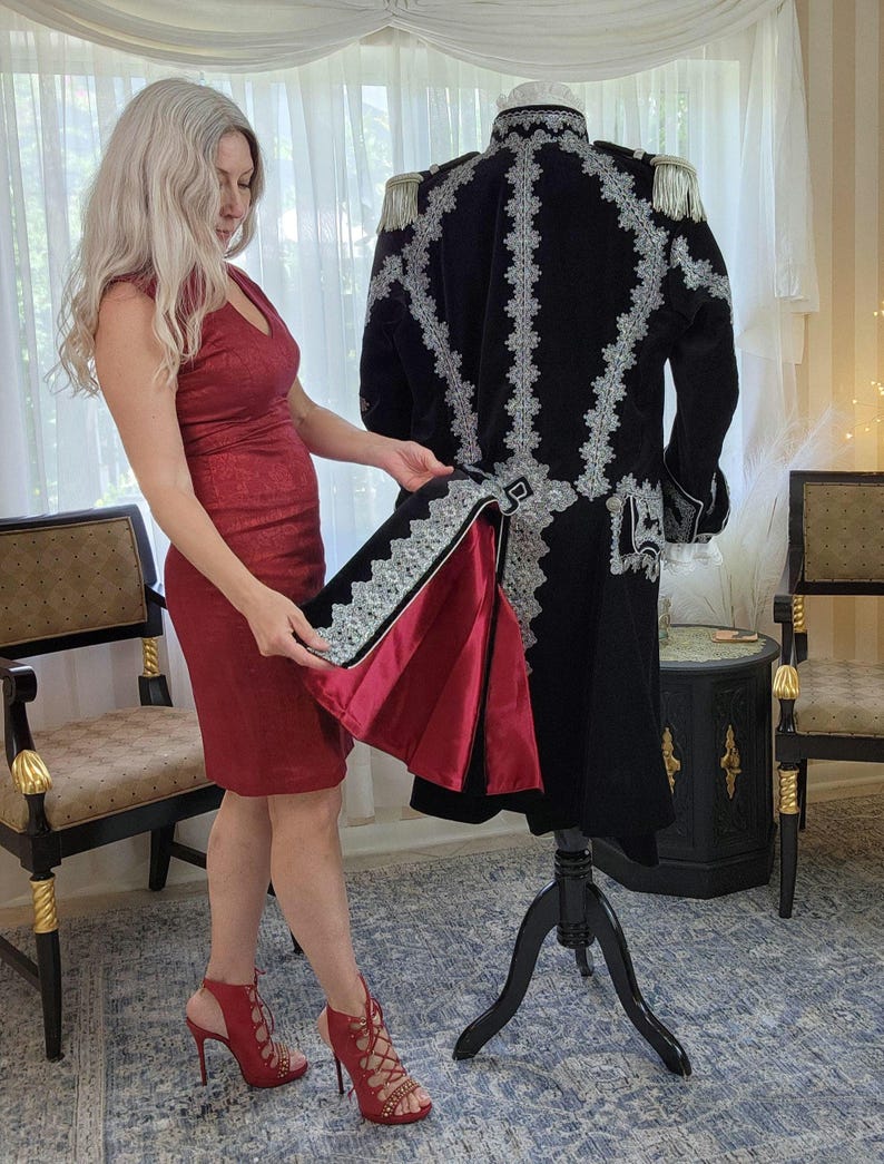 May include: A black military-style coat with intricate silver embroidery and epaulettes is displayed on a mannequin. The coat's interior lining is a deep red. The coat is a vintage or theatrical garment.