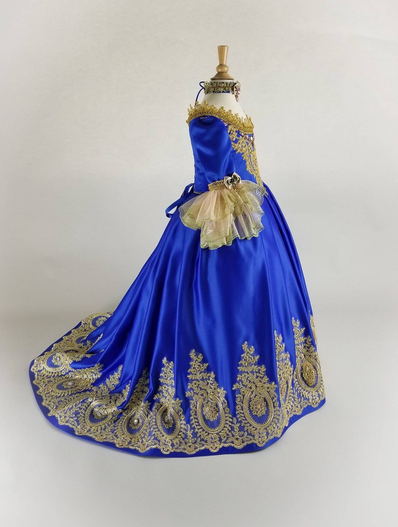 K&ouml;nnte beinhalten: Ein k&ouml;niglich blaues Satin-Ballkleid mit goldenen Spitzendetails und einer T&uuml;llr&uuml;sche am &Auml;rmel. Das Kleid hat ein tailliertes Oberteil und einen vollen Rock.