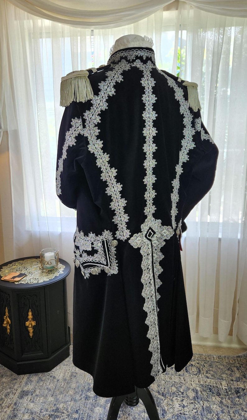 May include: A black velvet coat with white lace trim and epaulettes. The coat features intricate white lace detailing down the back and around the edges, with decorative buttons and a high collar. The coat is a formal garment.