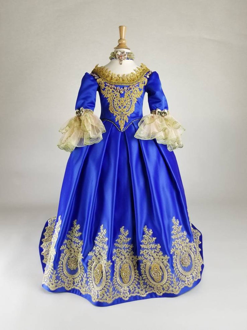 K&ouml;nnte beinhalten: Ein k&ouml;niglich blaues Satinkleid mit goldener Stickerei und Spitzendetails. Das Kleid hat ein tailliertes Oberteil und einen vollen Rock. Die &Auml;rmel sind lang und haben Spitzenr&uuml;sche an den &Auml;rmeln.