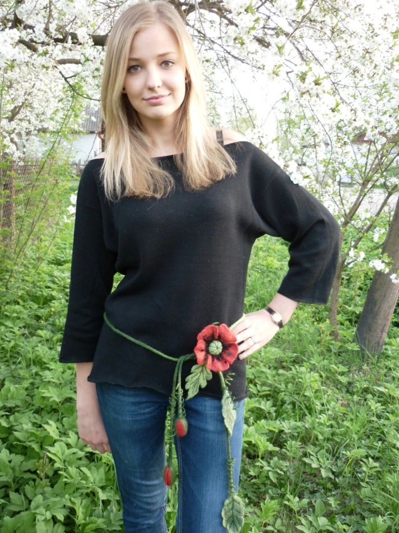 May include: A woman wearing a black sweater and blue jeans with a felt poppy flower belt.