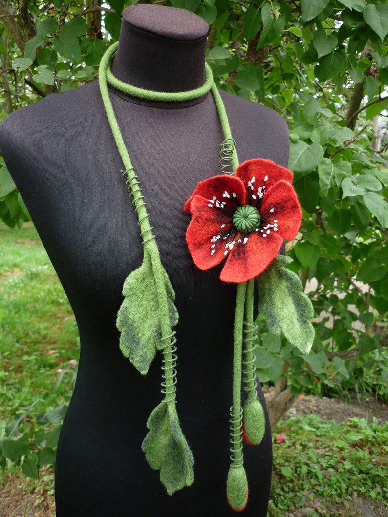 May include: A red felt poppy flower with white accents and green felt leaves hangs from a green felt cord with metal wire accents. The necklace is displayed on a black mannequin.