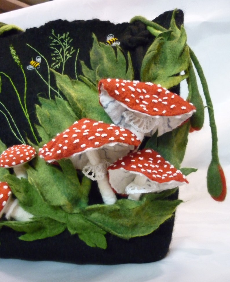 May include: A black felt bag with a drawstring closure, featuring embroidered red and white mushrooms with white lace accents and green leaves. Two yellow bees are embroidered on the bag.