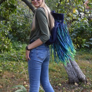 May include: A woman wearing blue jeans and a green long-sleeved shirt is wearing a blue backpack with long green, blue, and turquoise fringe.