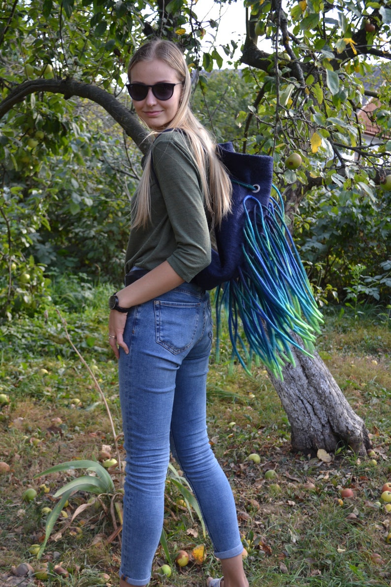 May include: A woman wearing blue jeans and a green long-sleeved shirt is wearing a blue backpack with long green, blue, and turquoise fringe.