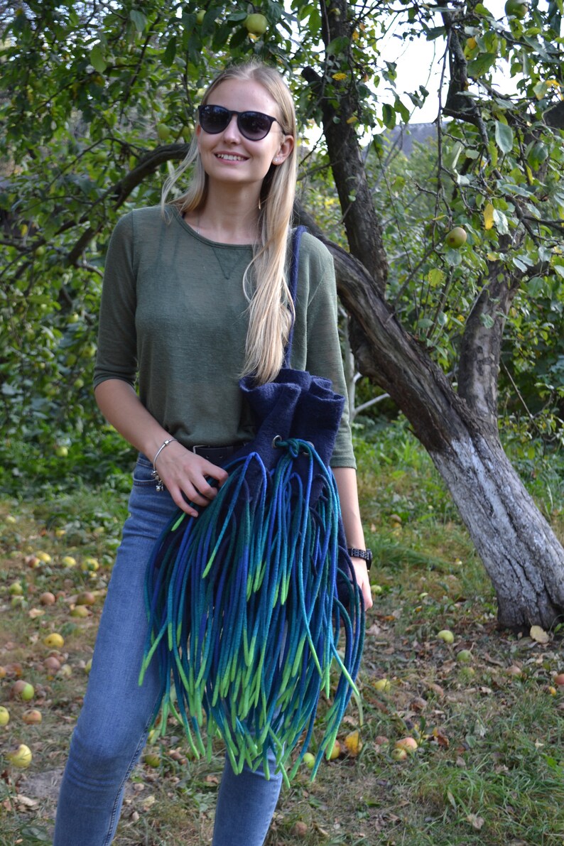 May include: A woman wearing sunglasses and a green shirt is holding a blue and green felted bag with long fringe. The bag is hanging from her shoulder. She is standing in front of a tree with green leaves and brown bark.
