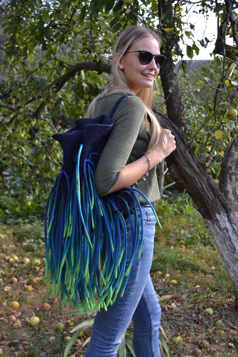 May include: A blue and green felted wool backpack with long fringe. The backpack is being worn by a person with blonde hair and sunglasses.