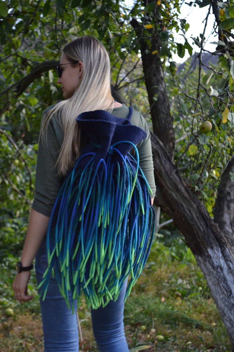 May include: A woman wearing blue jeans and a green shirt is carrying a blue and green felted backpack with long fringe.