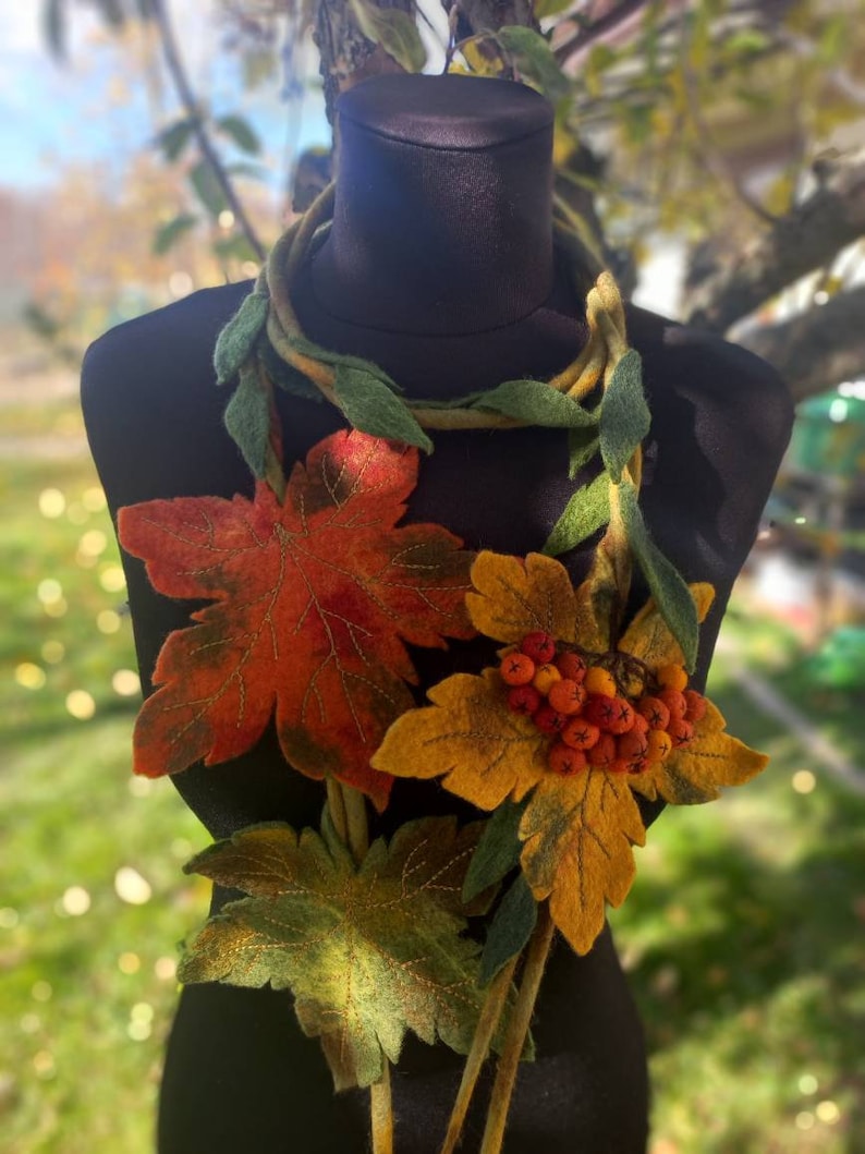 Puede incluir: Un collar de fieltro con hojas de oto&ntilde;o y bayas. El collar presenta una gran hoja de arce naranja y roja, una hoja amarilla con bayas naranjas y hojas verdes m&aacute;s peque&ntilde;as.