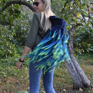 May include: A woman wearing blue jeans and a green shirt is carrying a blue and green fringed backpack.