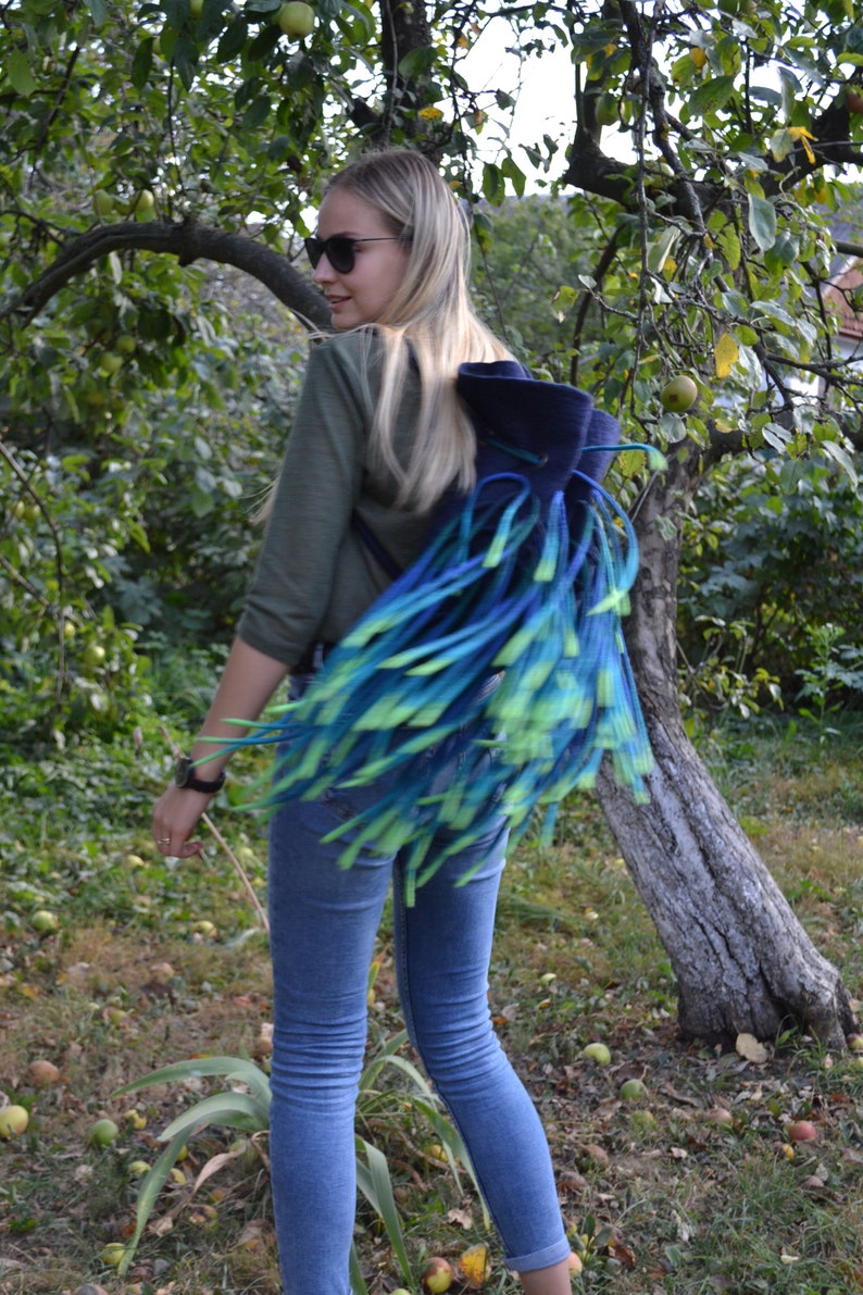May include: A woman wearing blue jeans and a green shirt is carrying a blue and green fringed backpack.