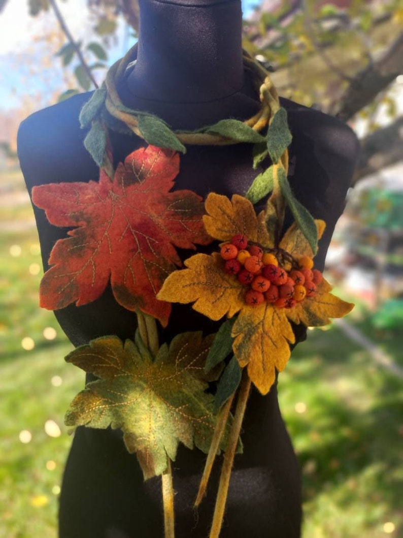Puede incluir: Un collar de fieltro con una hebra larga y fina de fieltro verde y marr&oacute;n. La hebra est&aacute; decorada con hojas de fieltro en tonos de verde, amarillo, naranja y rojo. Tambi&eacute;n hay peque&ntilde;as bayas rojas de fieltro en el collar.