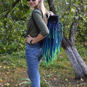 May include: A woman wearing sunglasses and a green long-sleeved shirt is standing in a garden. She is wearing blue jeans and carrying a blue and green fringed backpack.