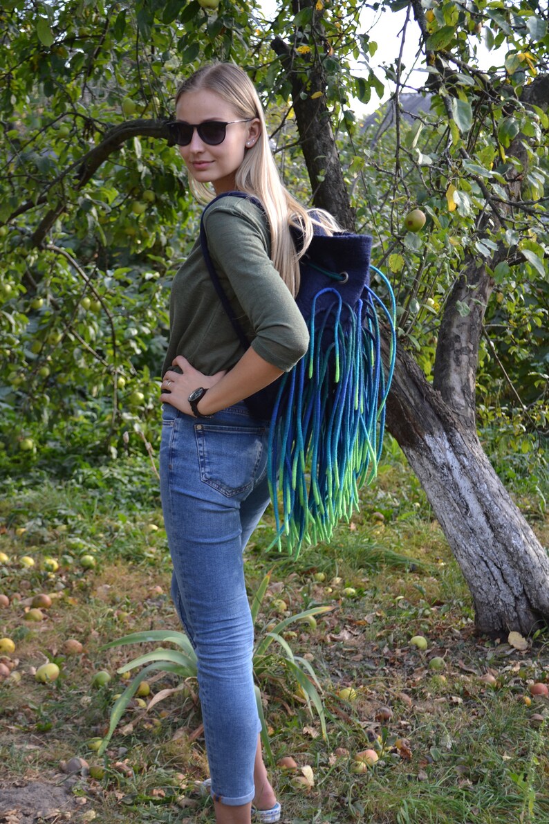 May include: A woman wearing sunglasses and a green long-sleeved shirt is standing in a garden. She is wearing blue jeans and carrying a blue and green fringed backpack.