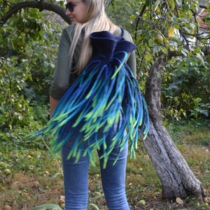 May include: A blue and green felted backpack with long fringe. The backpack is worn by a person with long blonde hair and sunglasses.