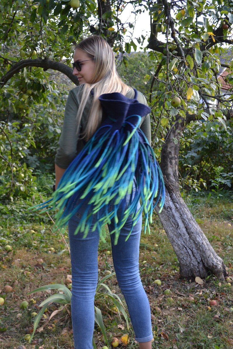 May include: A blue and green felted backpack with long fringe. The backpack is worn by a person with long blonde hair and sunglasses.