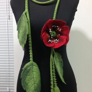 May include: A felt necklace with a red poppy flower and green leaves. The necklace is made of felt and is a unique and stylish accessory.